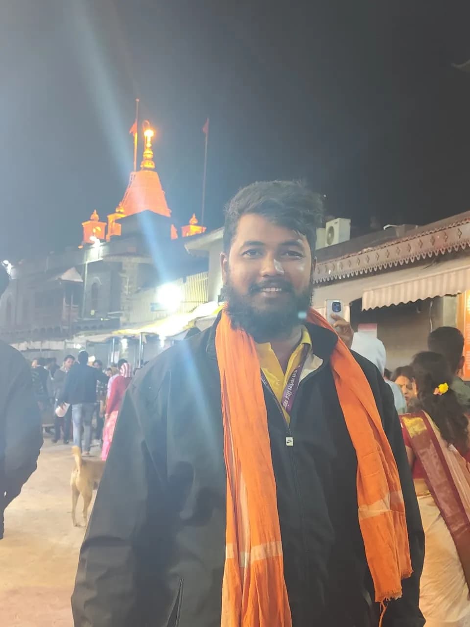 A devotee wearing a saffron scarf at a temple with a lit spire at night